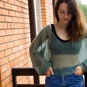 May include: A woman wearing a light green, sheer, knitted sweater with long sleeves and a loose fit. She is standing on a balcony with a brick wall in the background.