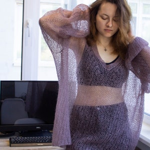 Könnte beinhalten: Eine Frau trägt einen durchsichtigen, helllila gestrickten Pullover mit V-Ausschnitt. Der Pullover ist locker geschnitten und hat ein zartes, offenmaschiges Muster.
