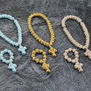 以下が含まれることがあります： 十字架が付いた結び目のついたお祈り用のビーズが3セット。1セットは薄い青色、1セットは金色、1セットはベージュです。