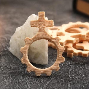 May include: A collection of light-colored wooden gear-shaped charms, each featuring a cross symbol at the top. The wooden pieces have a visible grain texture and are arranged in a cluster, with one piece standing upright against a white stone.