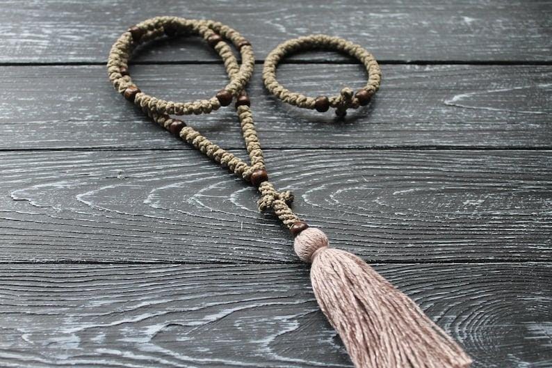 May include: A set of two brown and beige knotted prayer ropes with wooden beads and a tassel. The larger rope has a wooden cross at the end. The smaller rope is a bracelet.