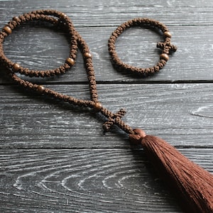 May include: A brown prayer rope with wooden beads and a tassel. The rope is knotted and has a cross at the end. There is a smaller brown bracelet with a cross in the image as well.