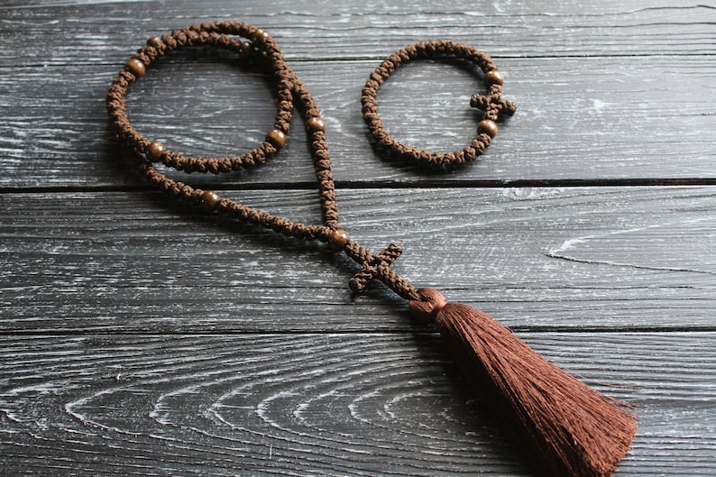 May include: A brown prayer rope with wooden beads and a tassel. The rope is knotted and has a cross at the end. There is a smaller brown bracelet with a cross in the image as well.