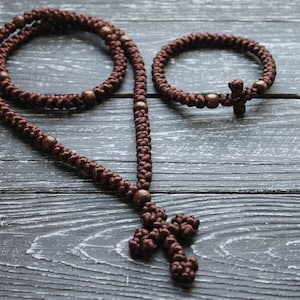 K&ouml;nnte beinhalten: Eine braune, gekn&uuml;pfte Schnurkette mit Holzperlen und einem Kreuzanh&auml;nger. Ein passendes braunes, gekn&uuml;pftes Schnurarmband mit Holzperlen und einem Kreuzanh&auml;nger ist ebenfalls abgebildet.