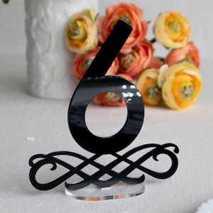 May include: A black acrylic table number six with an ornate base, set on a clear stand. The number is in front of a white vase with white flowers and orange and yellow flowers.