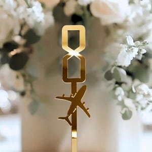 May include: A gold-colored table number holder with a rectangular top and an airplane silhouette. The holder is placed on a white surface, with a blurred floral arrangement in the background.