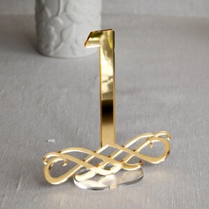 May include: A gold-colored table number one with an ornate base. The number is tall and slender, with a clear acrylic base. The background includes a white vase and a light gray tablecloth.