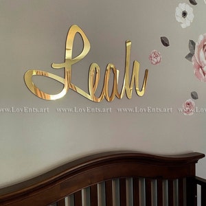 May include: Gold mirror acrylic name sign with the name "Leah" in a cursive font. The sign is mounted on a light beige wall with floral wall decals.