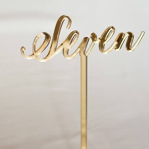 May include: Gold acrylic table number with the word "eleven" in a cursive font. The number is attached to a clear acrylic stand with a gold metal rod.