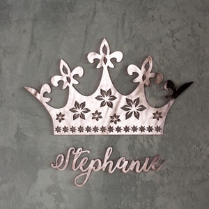 K&ouml;nnte beinhalten: Ros&eacute;goldene Kronen-Wanddekoration mit Blumen- und Sterndesign. Die Krone befindet sich &uuml;ber dem Namen "Stephanie" in Schreibschrift. Der Hintergrund ist eine strukturierte, graue Wand.