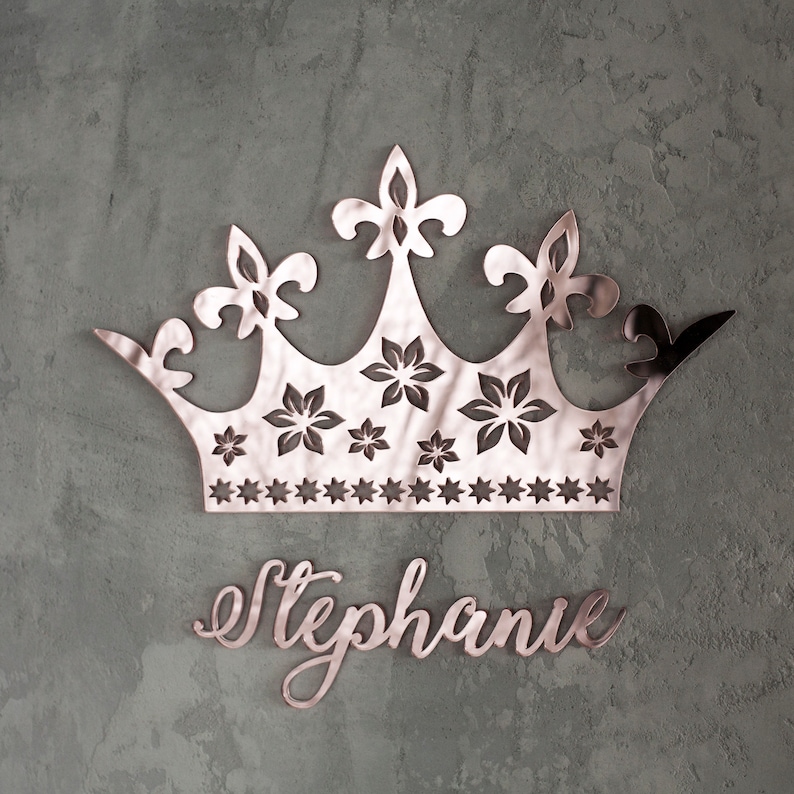 K&ouml;nnte beinhalten: Ros&eacute;goldene Kronen-Wanddekoration mit Blumen- und Sterndesign. Die Krone befindet sich &uuml;ber dem Namen "Stephanie" in Schreibschrift. Der Hintergrund ist eine strukturierte, graue Wand.