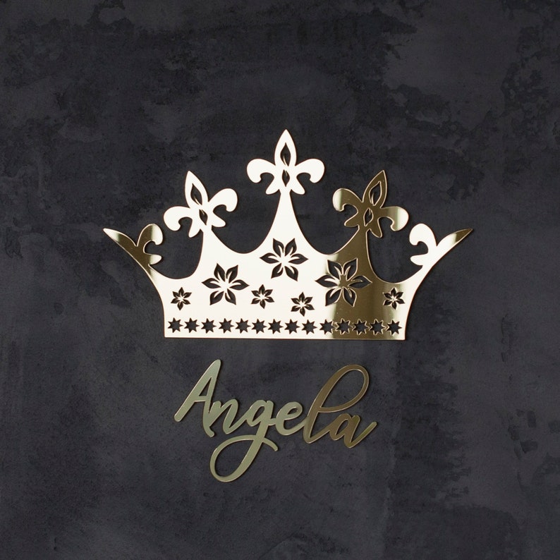 K&ouml;nnte beinhalten: Eine goldfarbene Krone mit floralen und sternf&ouml;rmigen Aussparungen und dem Namen "Angela" in Schreibschrift. Die Krone ist der Hauptfokus, vor einem dunklen, strukturierten Hintergrund. Das Design ist elegant und dekorativ.