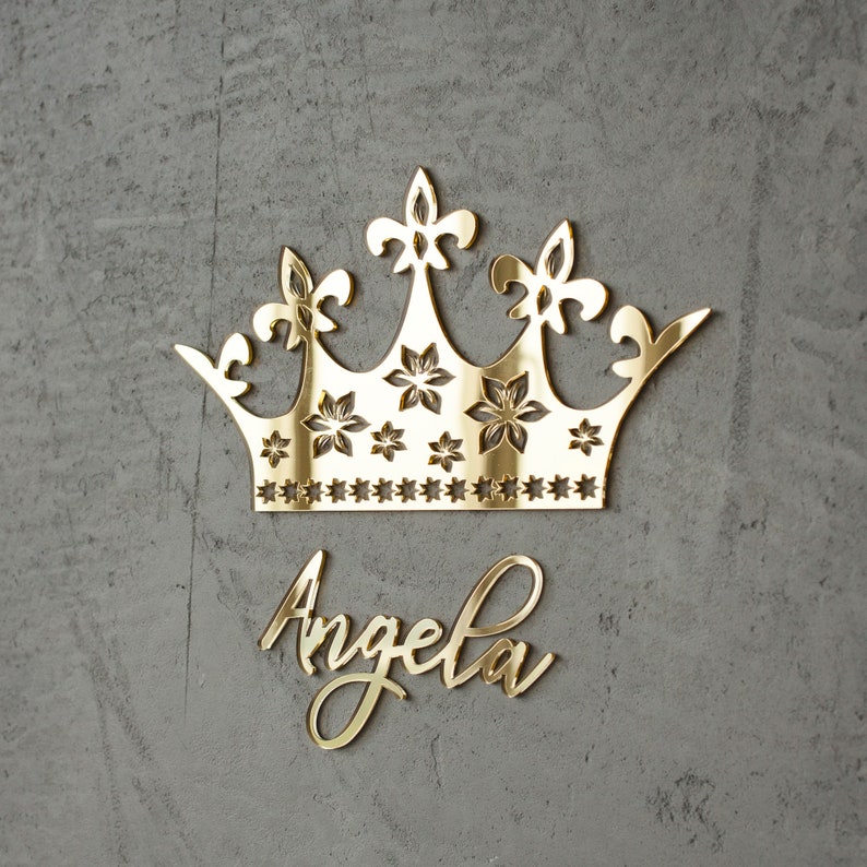 K&ouml;nnte beinhalten: Eine goldfarbene Krone mit floralen und Lilien-Details sowie dem Namen "Angela" in Schreibschrift. Die Krone ist ein dekorativer Artikel, m&ouml;glicherweise f&uuml;r eine Wand- oder Eventdekoration.