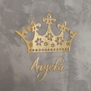 K&ouml;nnte beinhalten: Eine goldfarbene Krone und der Name "Angela" in Schreibschrift werden vor einem grau strukturierten Hintergrund dargestellt. Die Krone hat Fleur-de-Lis- und sternf&ouml;rmige Ausschnitte. Der Name ist in einer stilvollen, flie&szlig;enden Schrift.