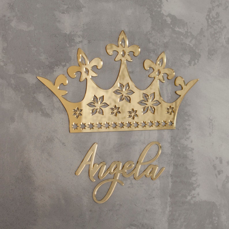 K&ouml;nnte beinhalten: Eine goldfarbene Krone und der Name "Angela" in Schreibschrift werden vor einem grau strukturierten Hintergrund dargestellt. Die Krone hat Fleur-de-Lis- und sternf&ouml;rmige Ausschnitte. Der Name ist in einer stilvollen, flie&szlig;enden Schrift.