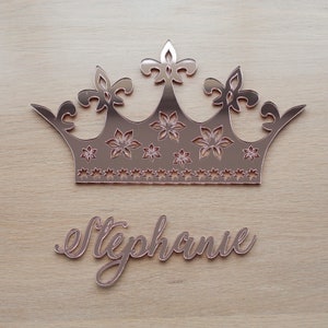 K&ouml;nnte beinhalten: Ros&eacute;goldfarbene, kronenf&ouml;rmige Wanddekoration mit floralen und sternf&ouml;rmigen Aussparungen sowie dem Namen "Stephanie" in Schreibschrift. Die Krone hat eine reflektierende Oberfl&auml;che und wird auf hellem Holzmaserungshintergrund angezeigt.