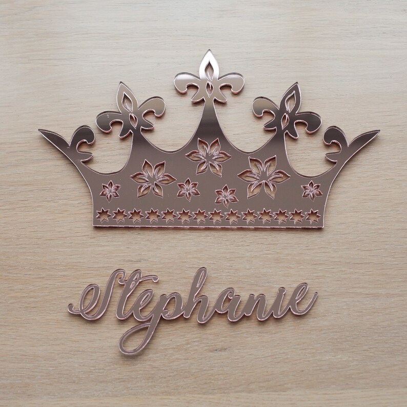 K&ouml;nnte beinhalten: Ros&eacute;goldfarbene, kronenf&ouml;rmige Wanddekoration mit floralen und sternf&ouml;rmigen Aussparungen sowie dem Namen "Stephanie" in Schreibschrift. Die Krone hat eine reflektierende Oberfl&auml;che und wird auf hellem Holzmaserungshintergrund angezeigt.