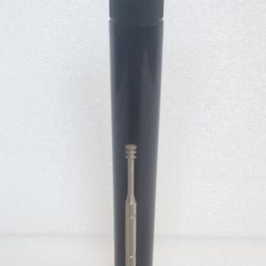May include: A tall, black cylindrical container with a lid. A silver-coloured, slender tool with a hook-like end is positioned next to the container. The tool features a textured grip section.