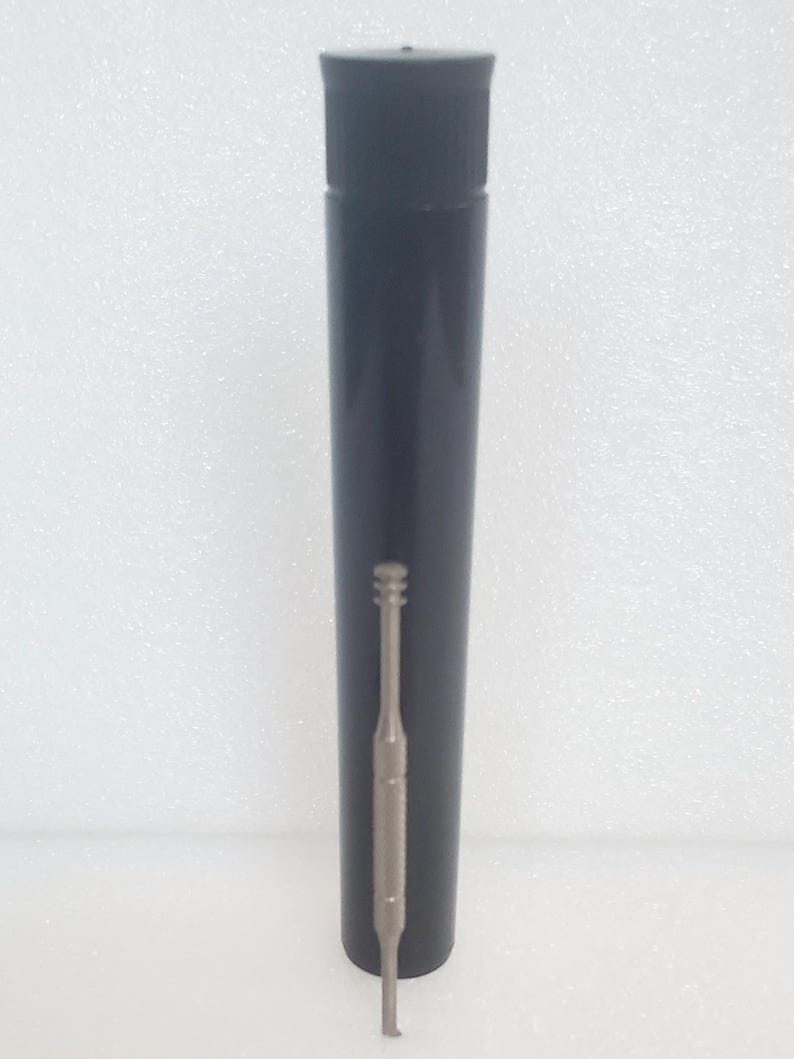 May include: A tall, black cylindrical container with a lid. A silver-coloured, slender tool with a hook-like end is positioned next to the container. The tool features a textured grip section.