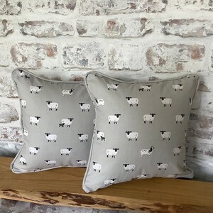 A gray throw pillow with a white border and a repeating pattern of black-faced sheep on a light gray background. Featuring the Sophie Allport Sheep fabric. The pillow is on a wood surface.