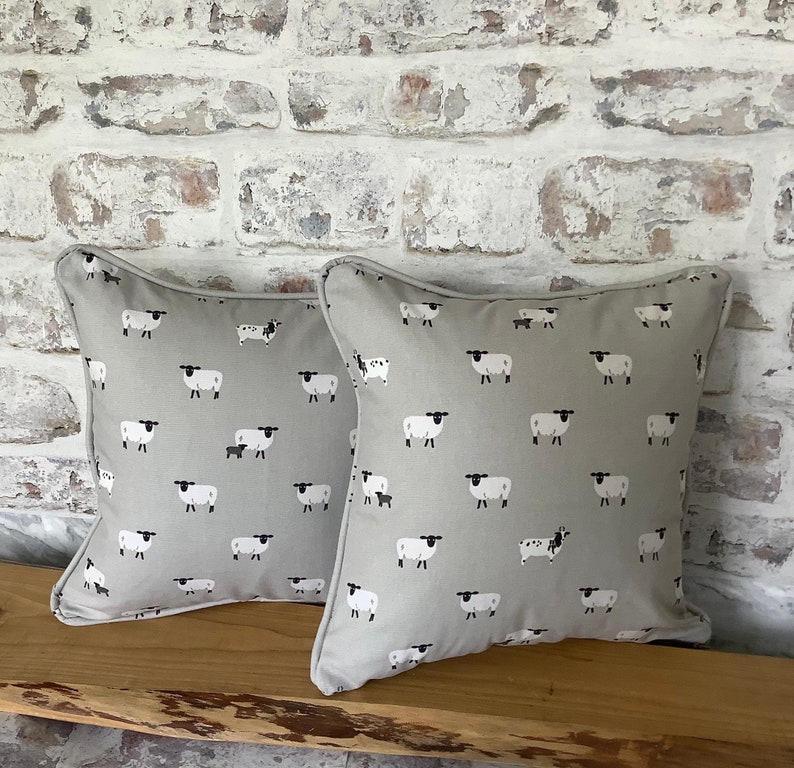 A gray throw pillow with a white border and a repeating pattern of black-faced sheep on a light gray background. Featuring the Sophie Allport Sheep fabric. The pillow is on a wood surface.