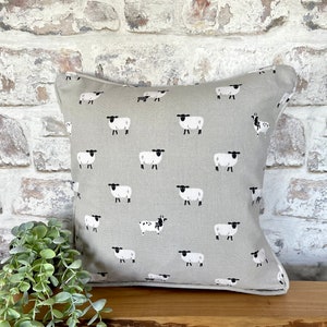 A gray throw pillow with a white border and a repeating pattern of black-faced sheep on a light gray background. Featuring the Sophie Allport Sheep fabric. The pillow is on a wood surface.