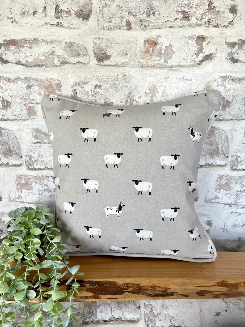A gray throw pillow with a white border and a repeating pattern of black-faced sheep on a light gray background. Featuring the Sophie Allport Sheep fabric. The pillow is on a wood surface.