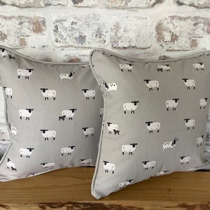 A gray throw pillow with a white border and a repeating pattern of black-faced sheep on a light gray background. Featuring the Sophie Allport Sheep fabric. The pillow is on a wood surface.