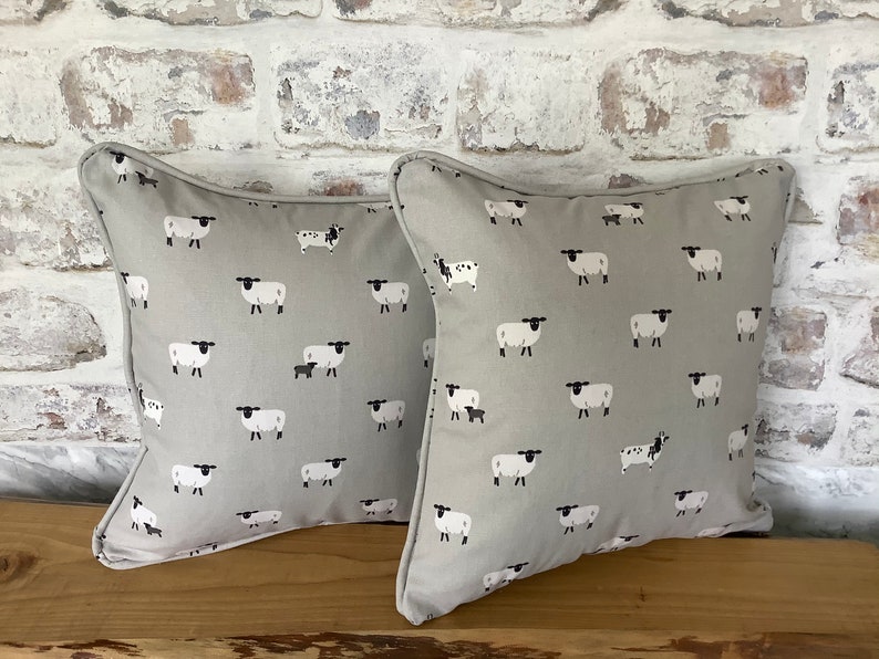 A gray throw pillow with a white border and a repeating pattern of black-faced sheep on a light gray background. Featuring the Sophie Allport Sheep fabric. The pillow is on a wood surface.