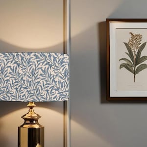 May include: A gold table lamp with a white lampshade featuring a blue floral pattern. The lampshade is rectangular and has a white background with blue leaves and branches. The lamp is on a white wall with a framed botanical print of a green plant with yellow flowers.