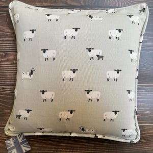 A gray throw pillow with a white border and a repeating pattern of black-faced sheep on a light gray background. Featuring the Sophie Allport Sheep fabric. The pillow is on a wood surface.