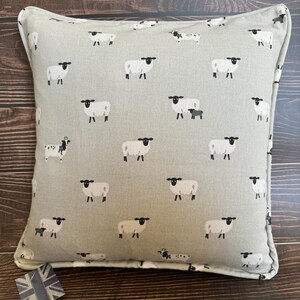 A gray throw pillow with a white border and a repeating pattern of black-faced sheep on a light gray background. Featuring the Sophie Allport Sheep fabric. The pillow is on a wood surface.