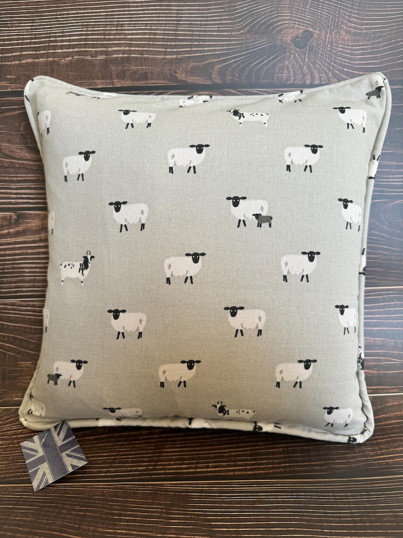 A gray throw pillow with a white border and a repeating pattern of black-faced sheep on a light gray background. Featuring the Sophie Allport Sheep fabric. The pillow is on a wood surface.