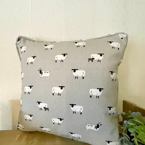 A gray throw pillow with a white border and a repeating pattern of black-faced sheep on a light gray background. Featuring the Sophie Allport Sheep fabric. The pillow is on a wood surface.