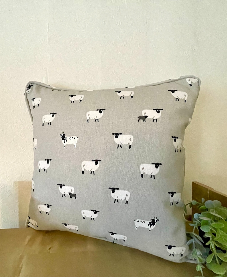 A gray throw pillow with a white border and a repeating pattern of black-faced sheep on a light gray background. Featuring the Sophie Allport Sheep fabric. The pillow is on a wood surface.