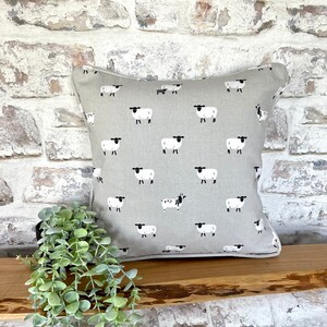 A gray throw pillow with a white border and a repeating pattern of black-faced sheep on a light gray background. Featuring the Sophie Allport Sheep fabric. The pillow is on a wood surface.