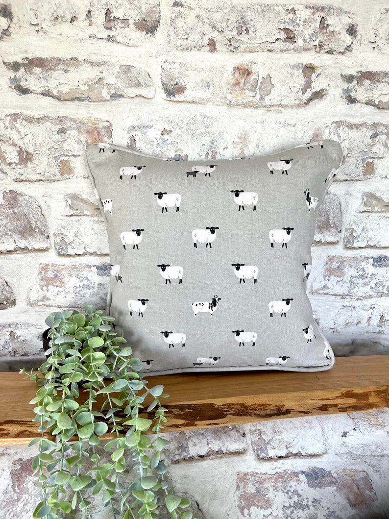 A gray throw pillow with a white border and a repeating pattern of black-faced sheep on a light gray background. Featuring the Sophie Allport Sheep fabric. The pillow is on a wood surface.
