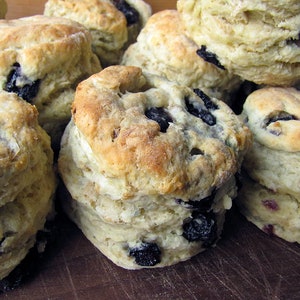 May include: A close-up of a pile of freshly baked blueberry biscuits. The biscuits are golden brown and have a flaky texture. They are arranged in a random pattern on a wooden surface.
