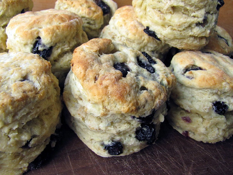 May include: A close-up of a pile of freshly baked blueberry biscuits. The biscuits are golden brown and have a flaky texture. They are arranged in a random pattern on a wooden surface.