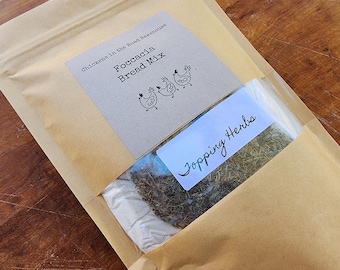 May include: A brown paper bag with a label that reads "Chickens in the Road Bakehouse Focaccia Bread Mix". The bag contains a clear plastic bag with dried herbs labeled "Topping Herbs".