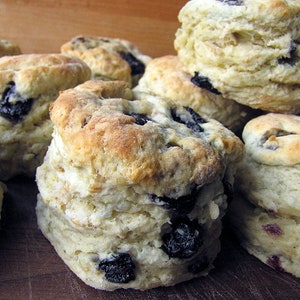 May include: A close-up of a pile of freshly baked blueberry biscuits. The biscuits are golden brown and have a flaky texture. They are topped with blueberries and are arranged on a wooden surface.
