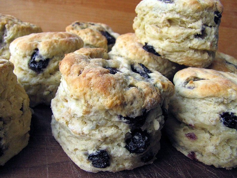 May include: A close-up of a pile of freshly baked blueberry biscuits. The biscuits are golden brown and have a flaky texture. They are topped with blueberries and are arranged on a wooden surface.