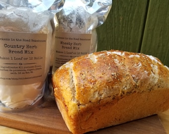 May include: Two bags of bread mix, one labelled "Country Herb Bread Mix" and the other labelled "Wheaty Herb Bread Mix", both from "Chickens in the Road Bakehouse".  Each bag makes one loaf or 12 rolls. A loaf of bread is in the foreground.