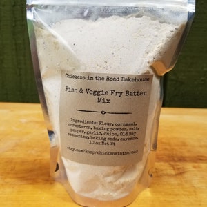 May include: A white bag of Fish & Veggie Fry Batter Mix from Chickens in the Road Bakehouse. The bag is labeled with the ingredients: Flour, cornmeal, cornstarch, baking powder, salt, pepper, garlic, onion, Old Bay seasoning, baking soda, and cayenne. The bag is 10 ounces.