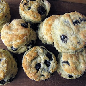 May include: A close-up of a group of blueberry biscuits on a wooden surface. The biscuits are light brown and have a slightly crispy texture. They are topped with blueberries and have a dusting of powdered sugar.
