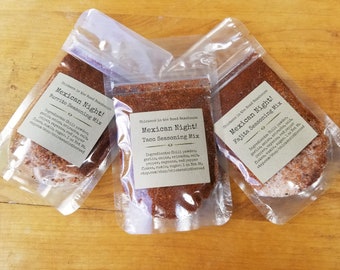May include: Three small clear plastic bags filled with red and brown spice blends. Each bag has a label that reads "Mexican Night!" with a different type of seasoning mix: Burrito, Taco, and Fajita. The labels also include the name "Chickens in the Road Roadhouse" and the ingredients for each blend.