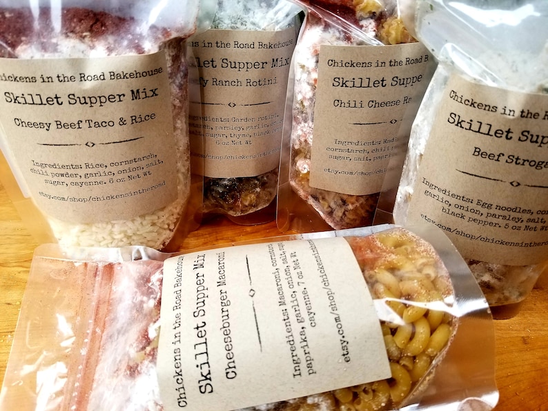 May include: Five clear plastic bags containing different types of skillet supper mixes. The bags are labeled with the name of the mix, the ingredients, and the net weight. The mixes include cheesy beef taco & rice, ranch rotini, chili cheese, beef stroganoff, and cheeseburger macaroni.
