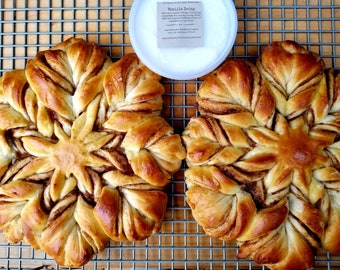 May include: Two golden brown snowflake-shaped cinnamon rolls on a wire rack. The rolls are topped with a vanilla icing.