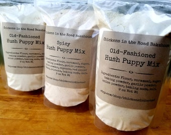May include: Three brown paper bags filled with a white powder mix. The bags are labelled "Chickens in the Road Bakehouse" and have the text "Old-Fashioned Hush Puppy Mix" and "Spicy Hush Puppy Mix". Each bag lists the ingredients: flour, cornmeal, sugar, baking powder, garlic powder, onion powder, baking soda, salt. The bags are 9 oz net weight.