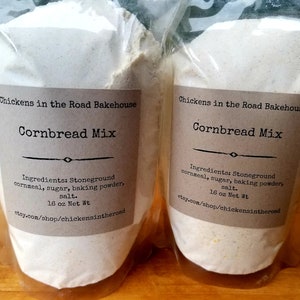 May include: Two clear plastic bags filled with a white cornbread mix. The bags have brown labels with the text "Chickens in the Road Bakehouse" and "Cornbread Mix". The labels also list the ingredients: stoneground cornmeal, sugar, baking powder, and salt. The bags are 16 ounces each.
